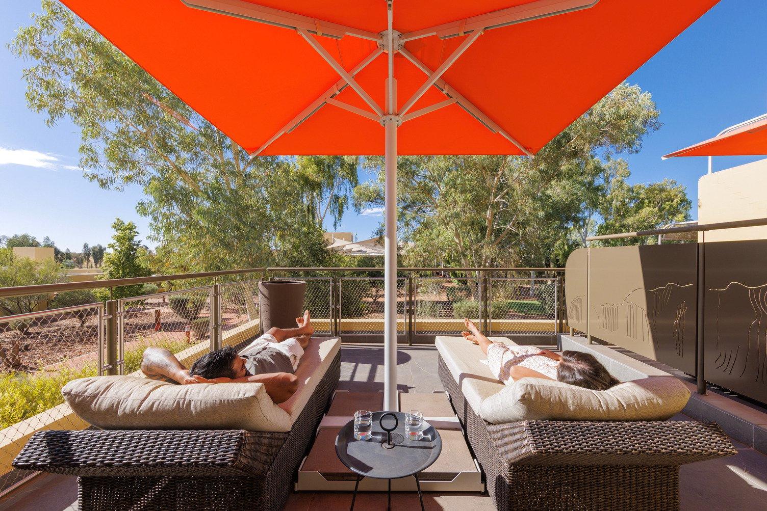 A couple lay on a sun lounge each with a large orange umbrella providing shade on the balcony