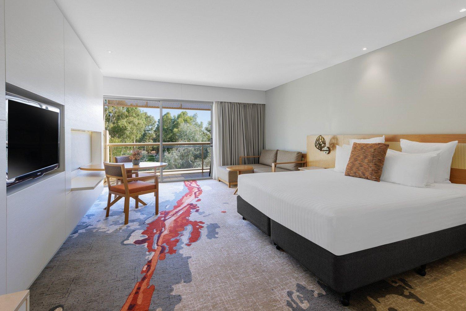 A king bed with white sheets, grey couch near the balcony with curtains open, table and chairs, and TV. 