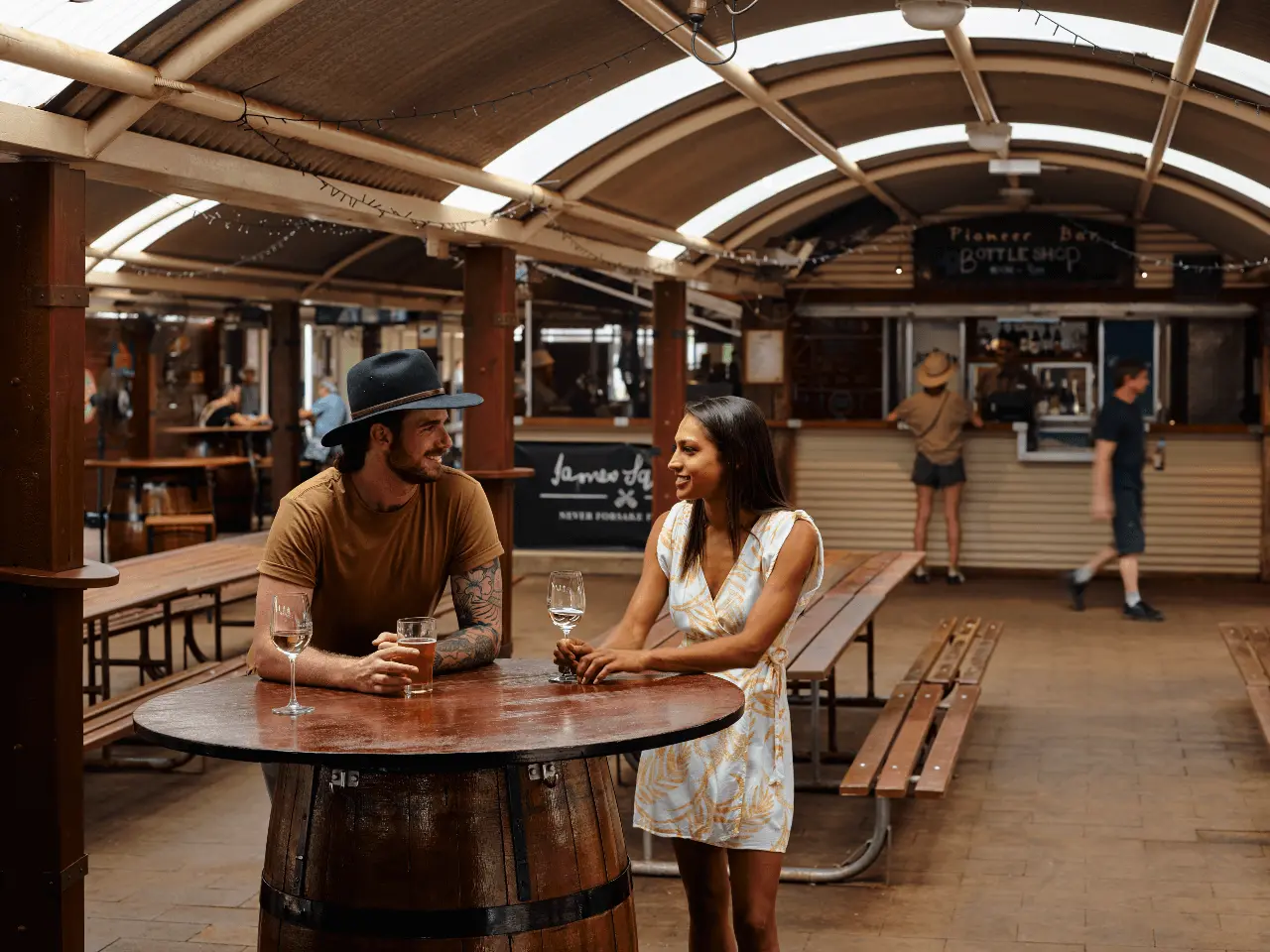 A couple standing at a table drinking a schooner of beer with people standing at the Outback Bottle Shop in the distance
