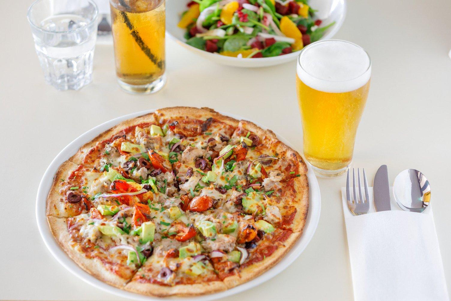 A plate of sliced Chicken Avocado pizza with a glass of beer, knife and fork and Beetroot and Fennel salad close by