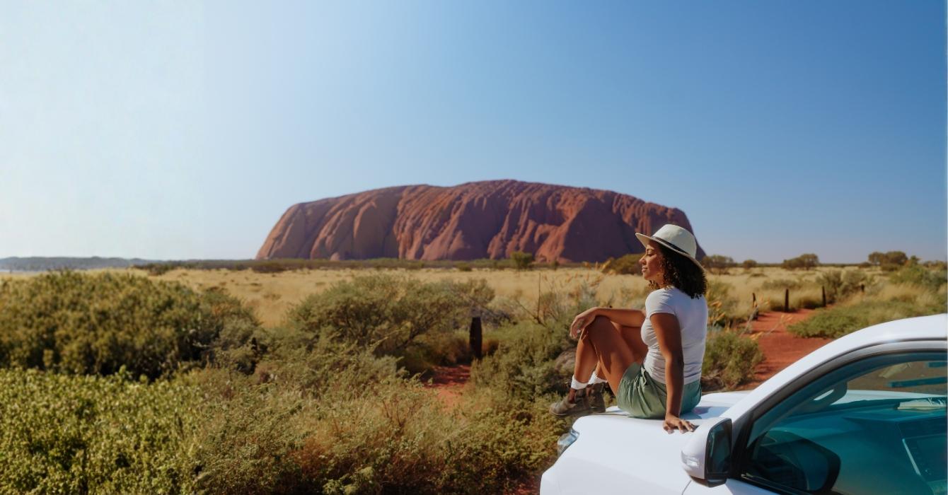 A girl sitting on top of a car with Uluru in the distance
