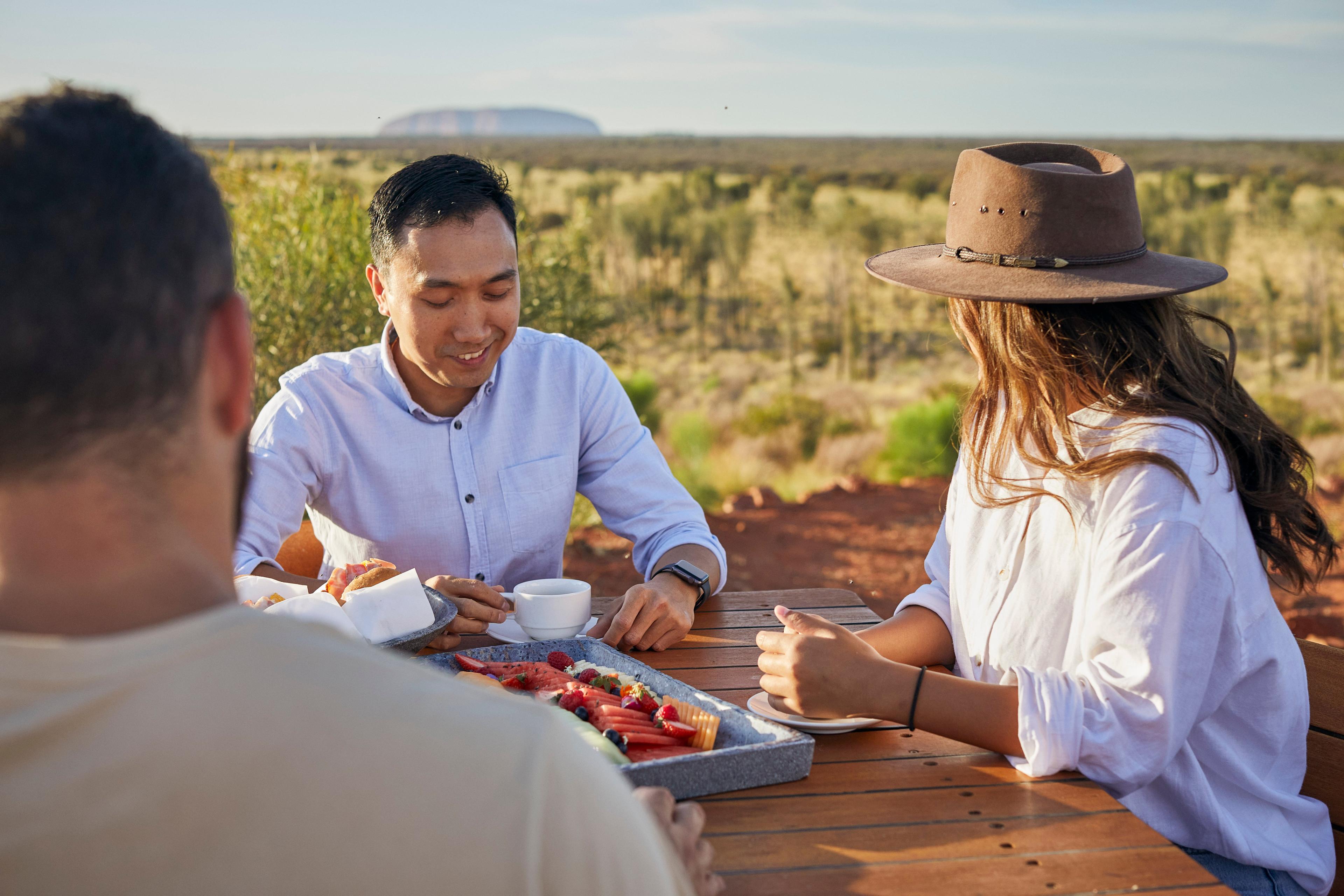 3 ppeople enjoying a breakfast platter and coffee with uluru and outback landscape in the backgroud